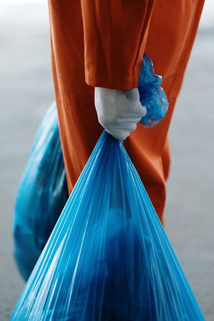 Close-up of a person wearing orange work overalls and a white glove, holding a tightly knotted blue plastic rubbish bag by the gathered top in their right hand. The bag appears semi-transparent with v